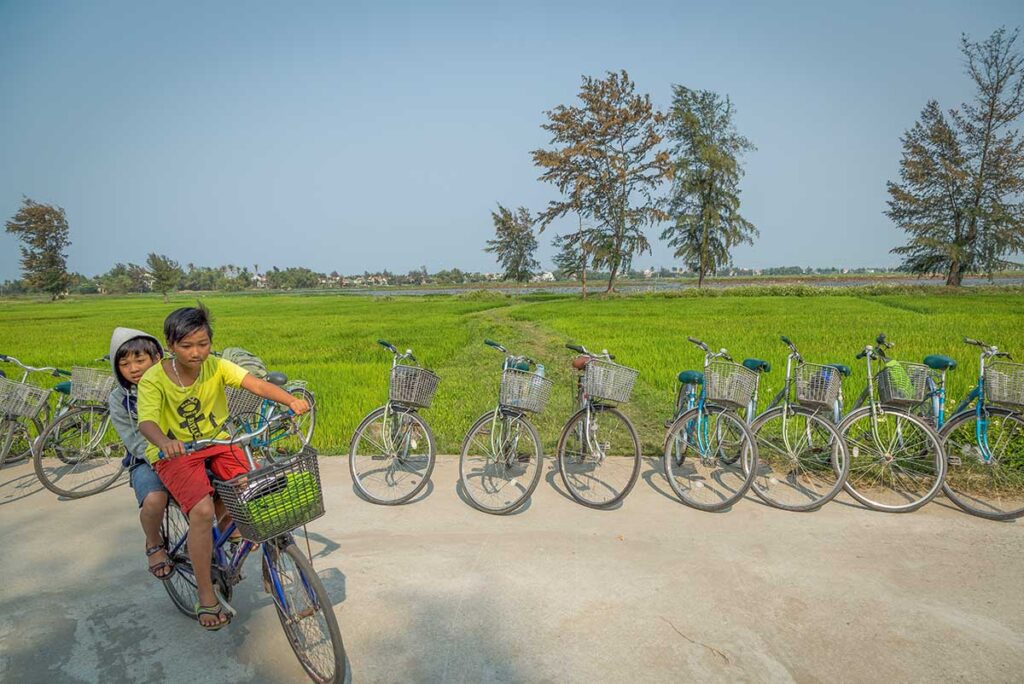 Local children cycling through green rice fields near Hoi An in March, when the weather turns warm and dryβideal for countryside bike tours and exploring village life.