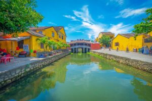 Bright sunny view of the Japanese Bridge in Hoi An in May, with clear skies and vibrant yellow houses reflecting the peak of the dry, warm season.