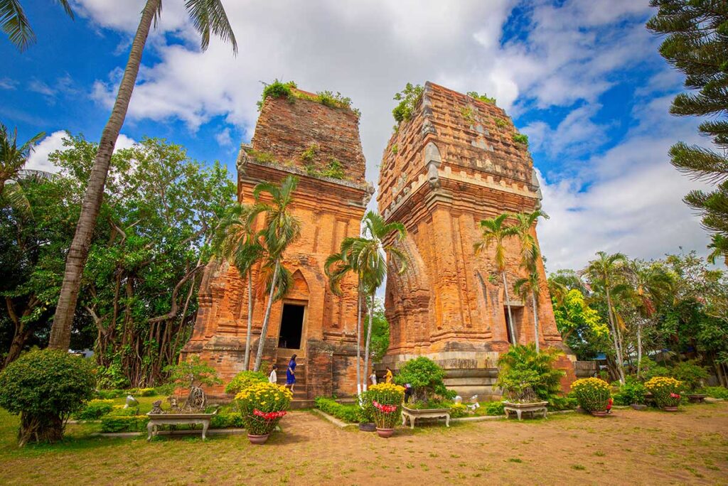 Front view of Cham Twin Towers in Quy Nhon city – Two red-brick towers standing side by side in a garden with palm trees.