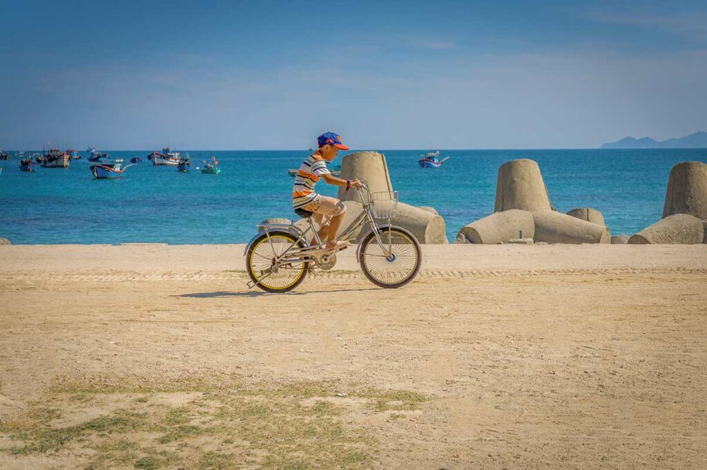 Local boy riding a bicycle along the seafront of Cu Lao Xanh Island, with blue fishing boats anchored in the background.