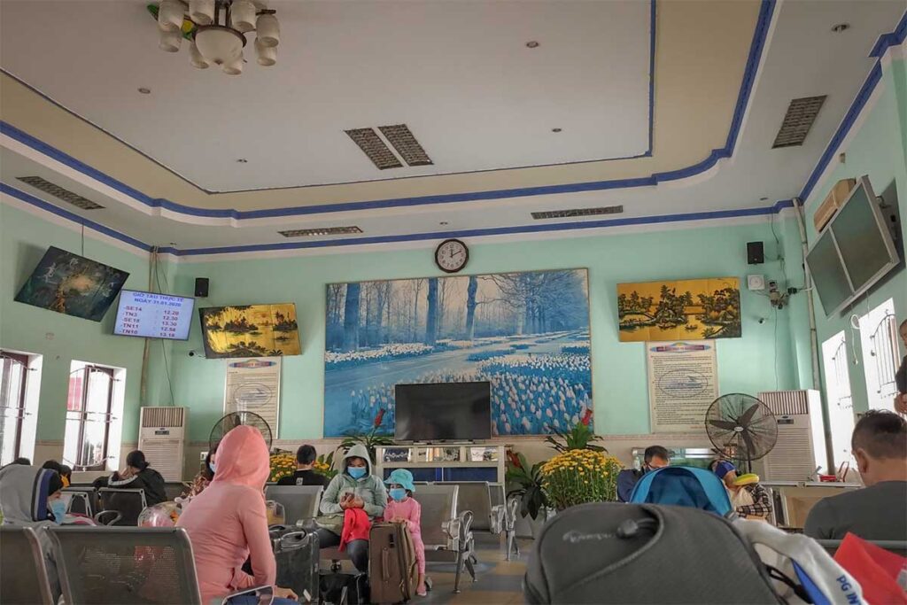 Waiting hall of Diêu Trì Train Station in Quy Nhon – passengers sitting in the modest waiting area with fans, digital schedule screens, and local artwork on the walls.