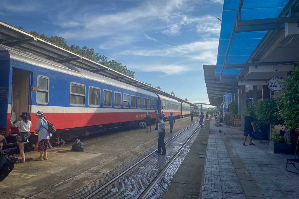 Platform at Diêu Trì Train Station – travelers boarding a north–south Reunification Express train under the covered platform on a sunny day.