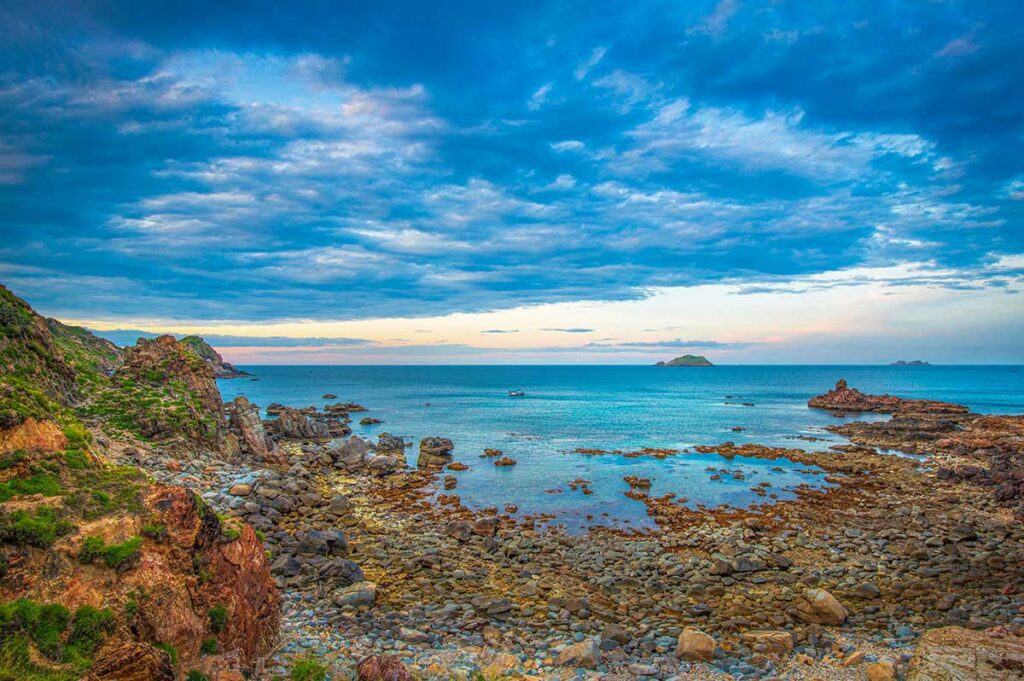 Rocky cove at Eo Gió with layered cliffs and calm turquoise water under a cloudy sky