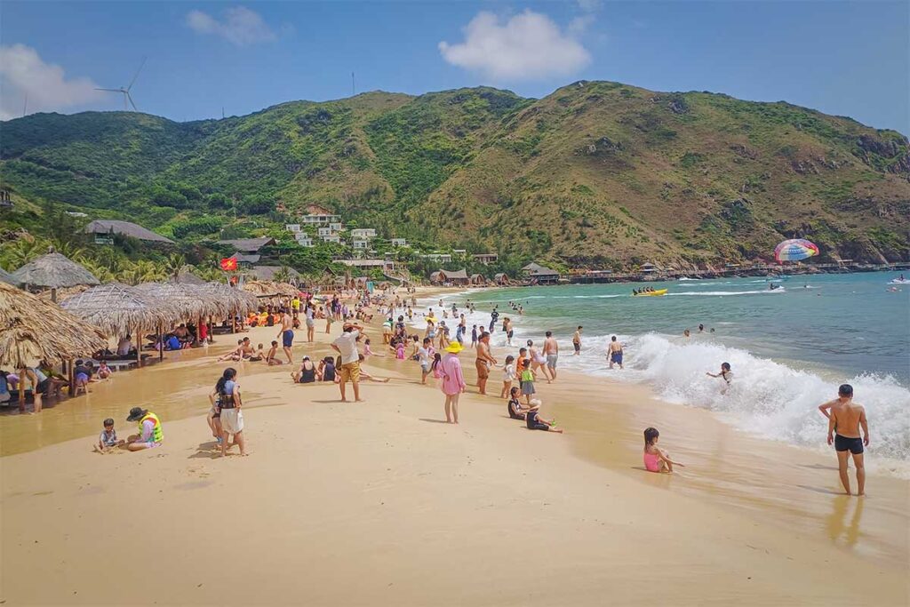 Crowded shoreline at Ky Co Beach – Tourists enjoying swimming and relaxing on the sandy beach, with green hills and wind turbines visible behind.
