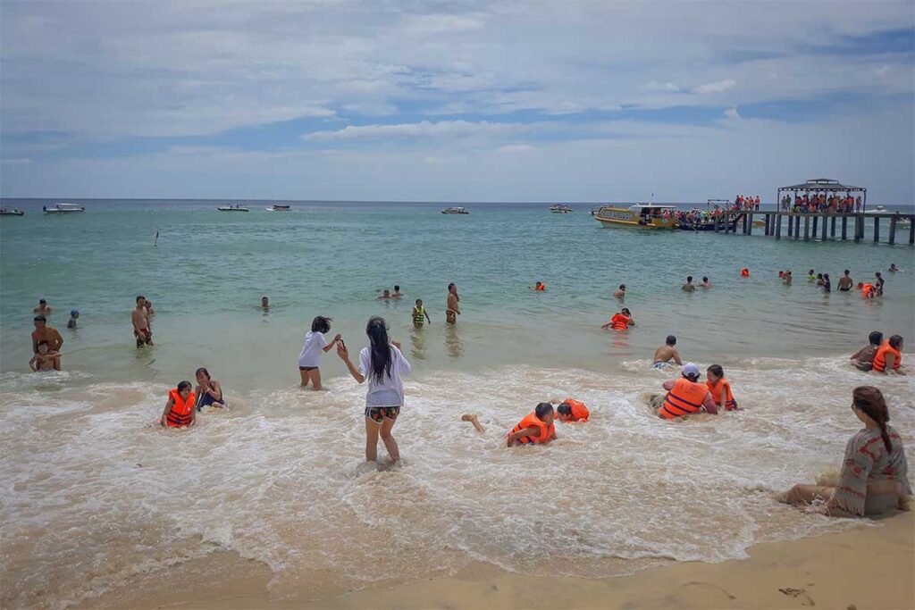 Families swimming at Ky Co Beach – Children and adults in life jackets playing in the surf near the pier and tour boats.