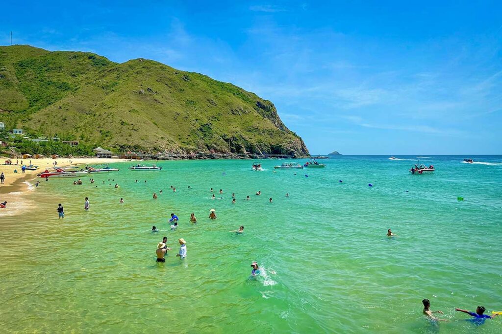 Crowds of tourists swimming and playing in the shallow turquoise water of Ky Co Beach in Quy Nhon, with speedboats anchored along the shore and green hills in the background.