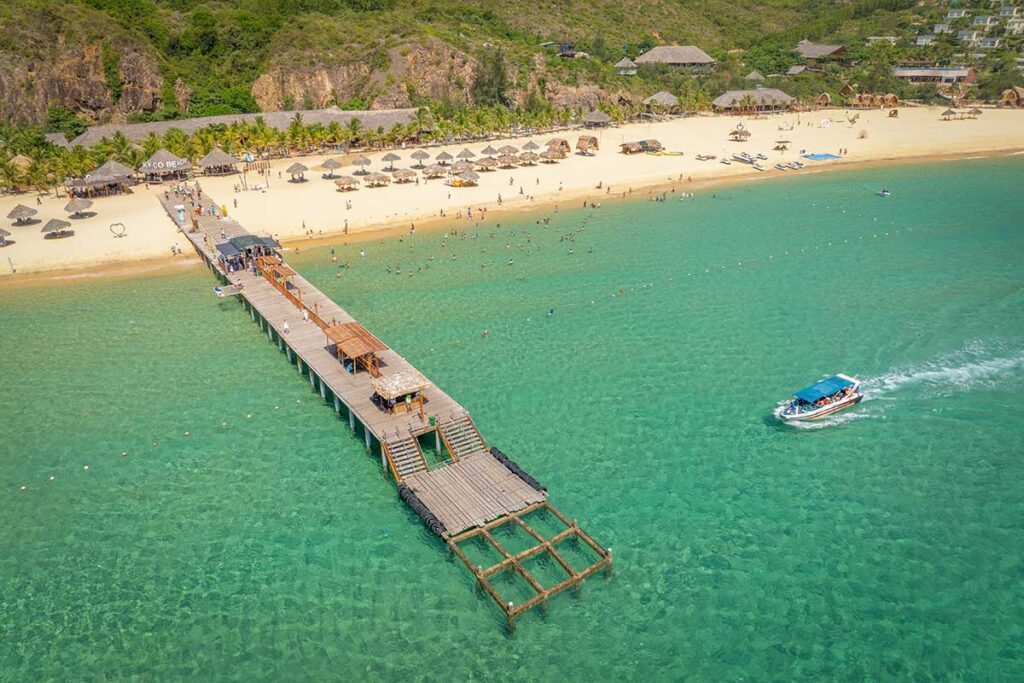 Aerial view of the wooden jetty at Ky Co Beach in Quy Nhon, with a speedboat cruising in crystal-clear water, tourists swimming nearby, and rows of thatched umbrellas on the sandy beach.