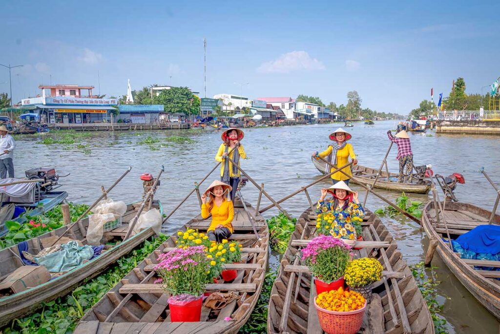 Women selling bright chrysanthemums from wooden boats on a sunny river, showing the festive vibe and dry, comfortable weather of the Mekong Delta in February.