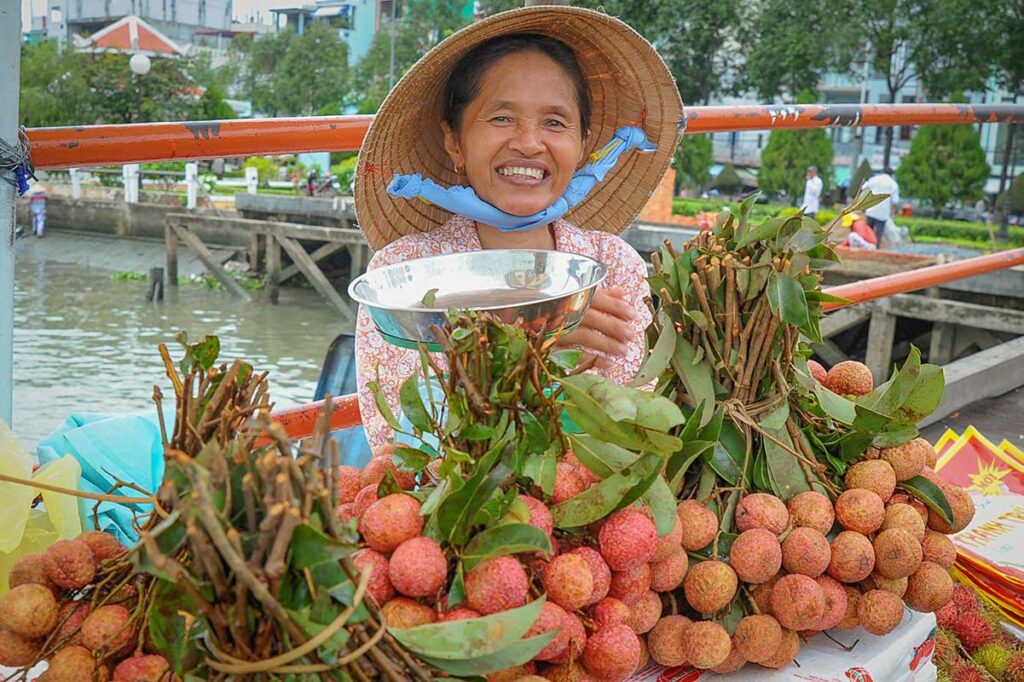 Smiling vendor weighing bunches of fresh lychees at a riverside stall, showing peak fruit harvest and humid rainy-season weather in the Mekong Delta in June.