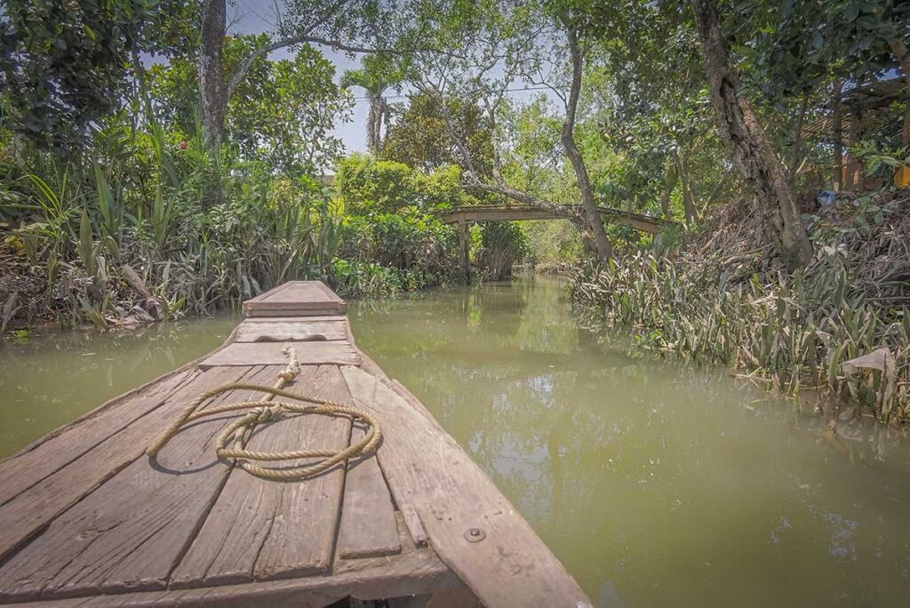 Wooden sampan gliding along a shaded canal beneath a small bridge, reflecting the clear, calm conditions of the Mekong Delta in March during the dry season.