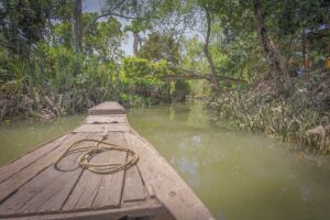 Wooden sampan gliding along a shaded canal beneath a small bridge, reflecting the clear, calm conditions of the Mekong Delta in March during the dry season.