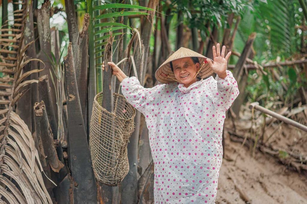 Smiling woman in conical hat and raincoat waving from riverside vegetation during a shower, reflecting the spirit of the Mekong Delta in rainy season.