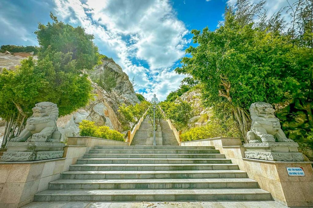 Long stone stairway to Ong Nui Temple’s seated Buddha, framed by lion statues and rocky cliffs.