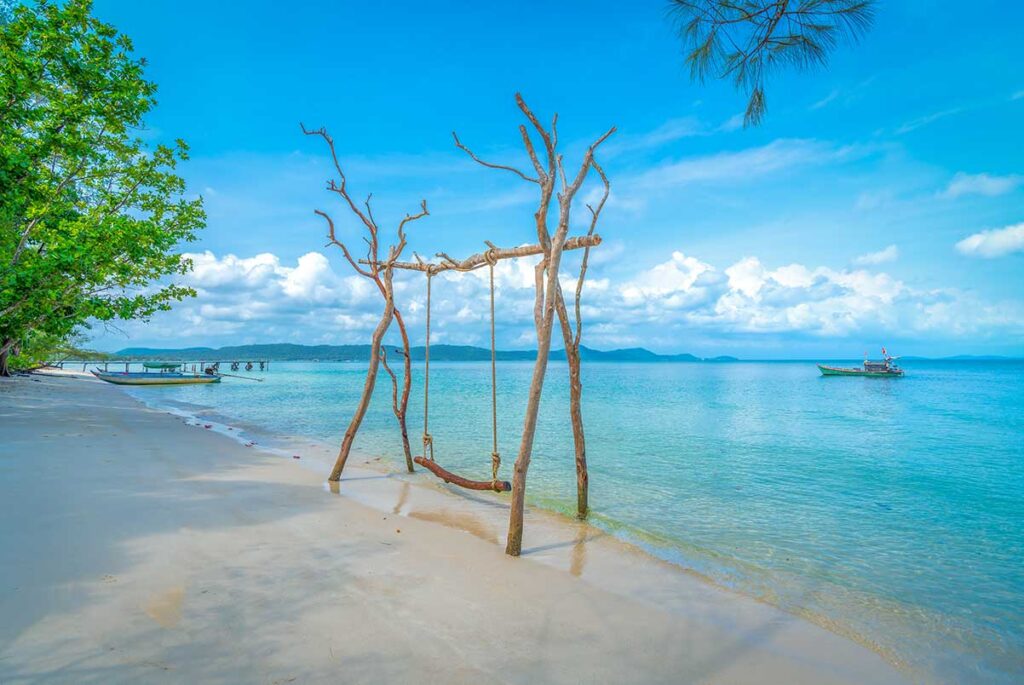Empty tropical beach with wooden swing, clear turquoise water, and fishing boat in the distance, showcasing the best time to visit Phu Quoc.