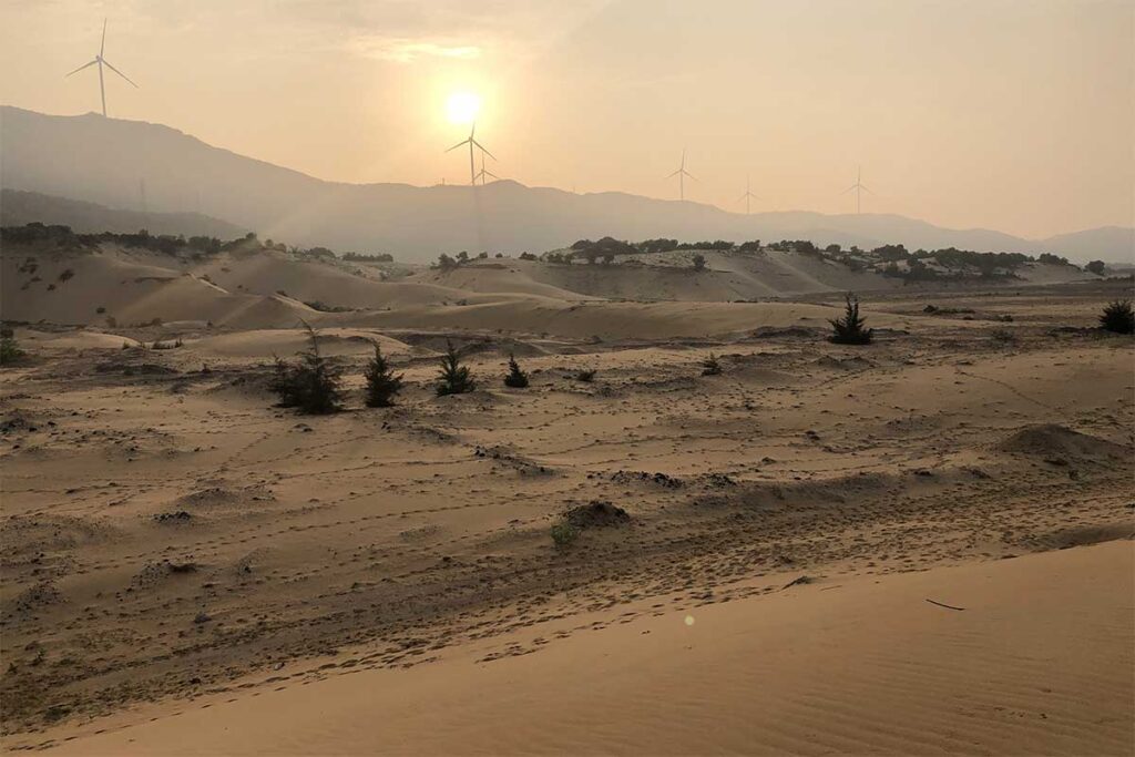 Golden sands of Phuong Mai at sunset with silhouettes of wind turbines on the ridge, blending renewable energy projects with natural landscapes