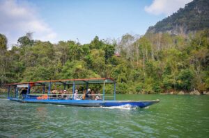 Travelers cruising on a traditional boat across the green waters of Ba Be Lake, with forested mountains in the background on a Ba Be Lake tour.