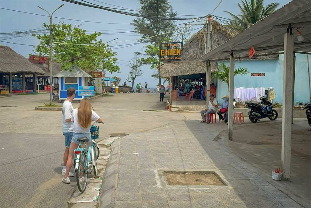 Travelers cycling through An Bang Beach village area near Hoi An, heading toward local seafood restaurants by the shore.
