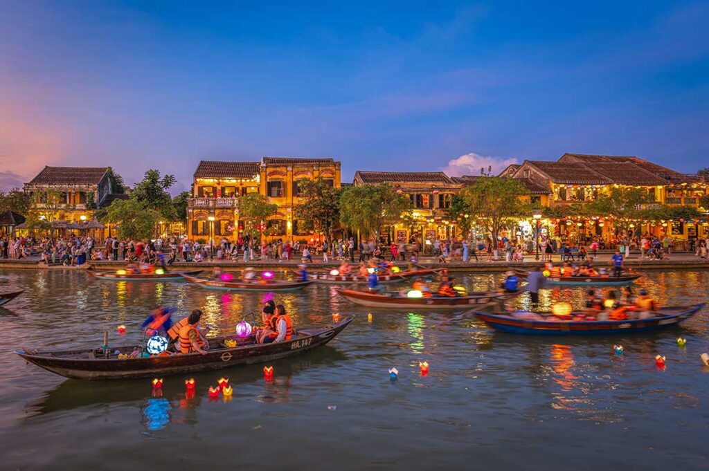 Evening boat ride on the river in Hoi An Ancient Town with visitors releasing colorful lanterns into the water under glowing street lights.