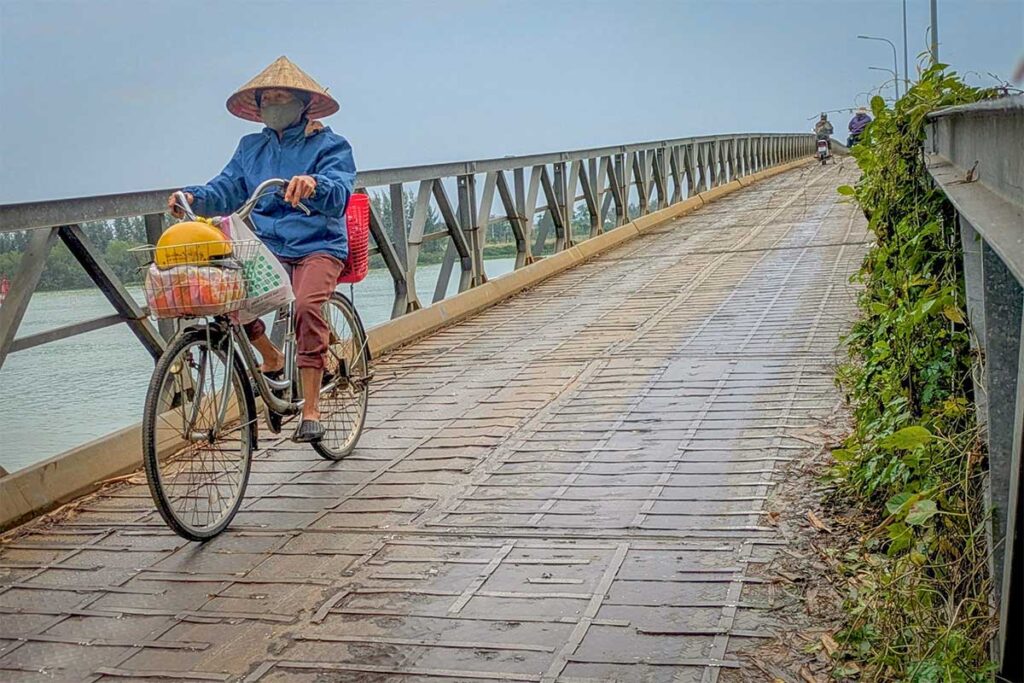 Cyclist crossing Cam Kim Bridge – Local woman in conical hat riding a bicycle across Cam Kim Bridge with baskets of produce, linking Hoi An to Cam Kim Island.