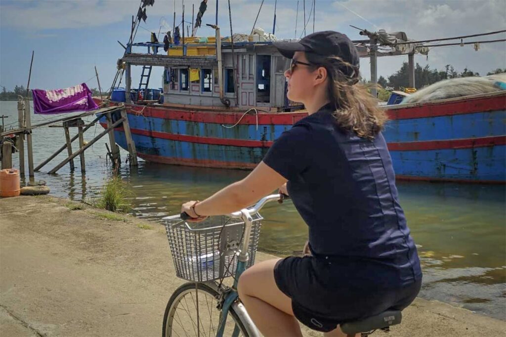 Tourist cycling along Cam Kim Island’s quiet rural path, passing traditional fishing boats and riverside scenery near Hoi An.