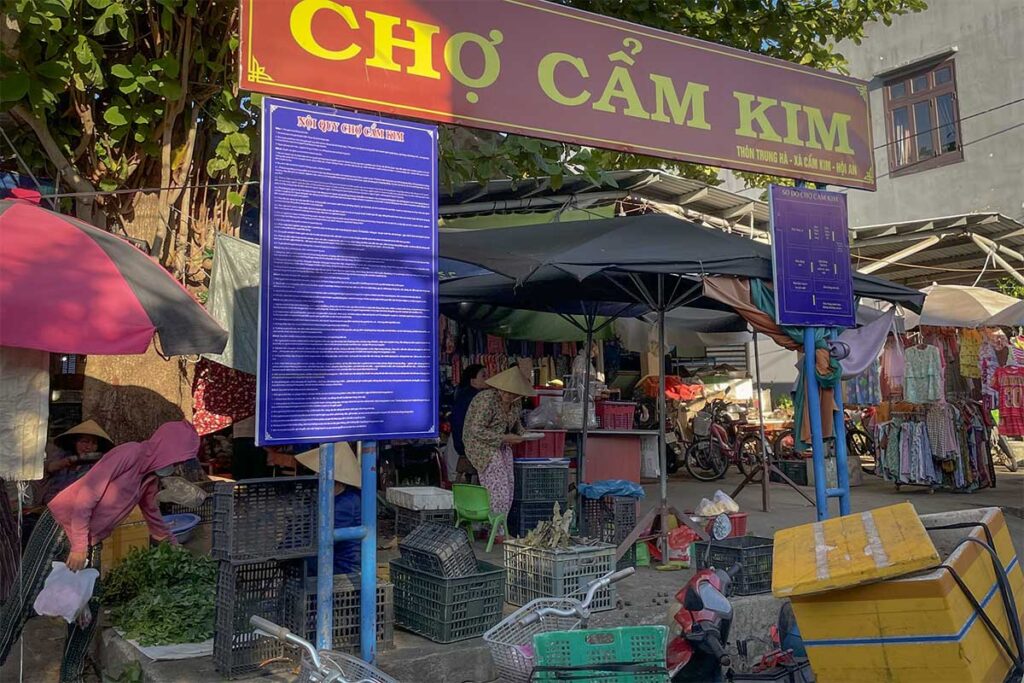 Entrance to Cam Kim Market in Hoi An with stalls selling vegetables, fruit, and daily essentials under shaded tarps