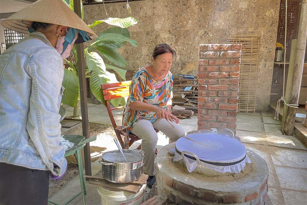 Elderly woman making traditional rice paper over a steaming clay stove in Cam Kim Village, Hoi An countryside.
