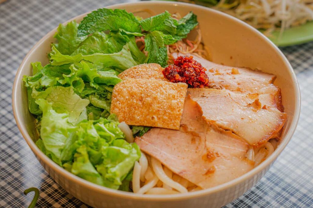 Authentic cao lầu noodles in Hoi An, topped with slices of pork, crispy rice crackers, fresh herbs, and chili paste, a signature local dish of the Ancient Town.