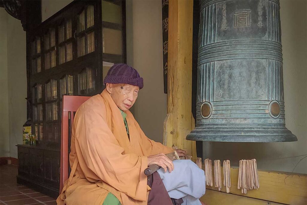 Elderly monk sitting beside ancient bronze bell at Chuc Thanh Pagoda in Hoi An – capturing spiritual life and Buddhist tradition in Vietnam.