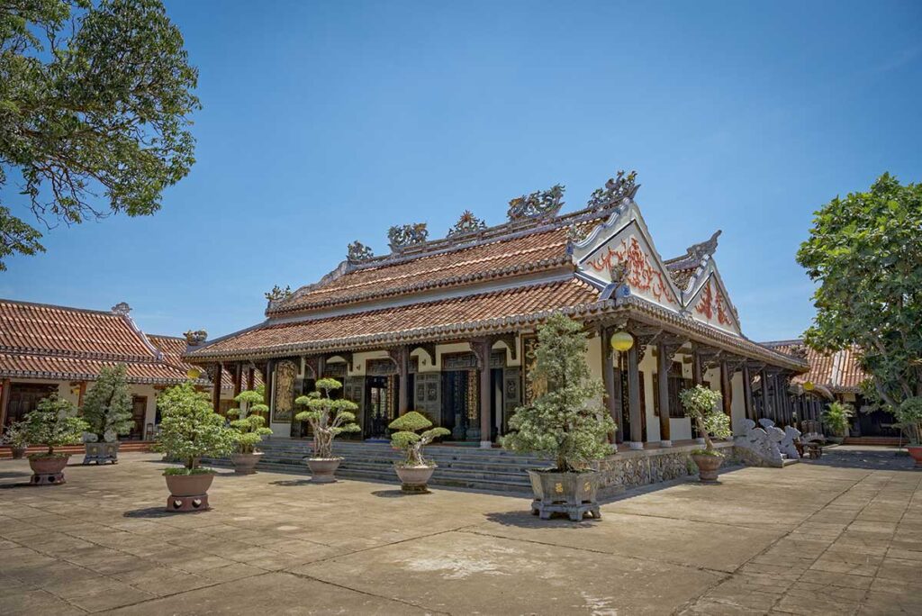Exterior courtyard of Chuc Thanh Pagoda Hoi An, Vietnam’s oldest Buddhist temple – tiled roofs and bonsai trees under clear blue sky.