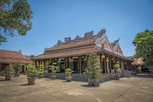 Exterior courtyard of Chuc Thanh Pagoda Hoi An, Vietnam’s oldest Buddhist temple – tiled roofs and bonsai trees under clear blue sky.