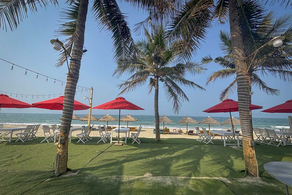 Beachfront restaurant with red umbrellas and palm trees at Cua Dai Beach Hoi An, offering sea views and relaxed dining.