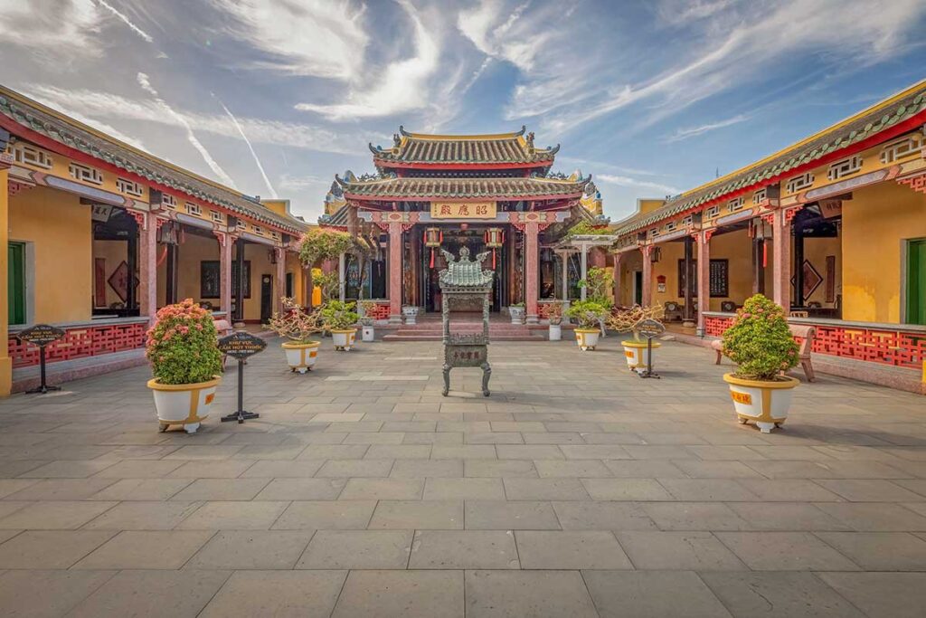 Open courtyard of Hai Nam Assembly Hall in Hoi An, bonsai planters and a central incense burner leading to the ornate main shrine.