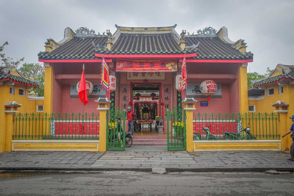 Street view of Hai Nam Assembly Hall Hoi An with old woman cycling past the entrance – authentic cultural scene combining local life and historic architecture.
