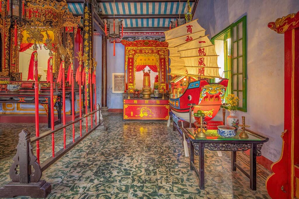 Colorful interior of Hai Nam Assembly Hall Hoi An with a wooden altar and red sailing boat model – richly decorated temple space honoring merchant traditions.