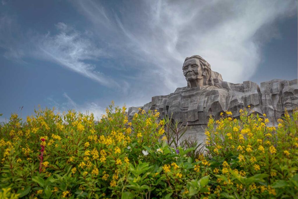Heroic Mother Thu Statue viewed behind yellow flowers under blue sky in Quang Nam Province.