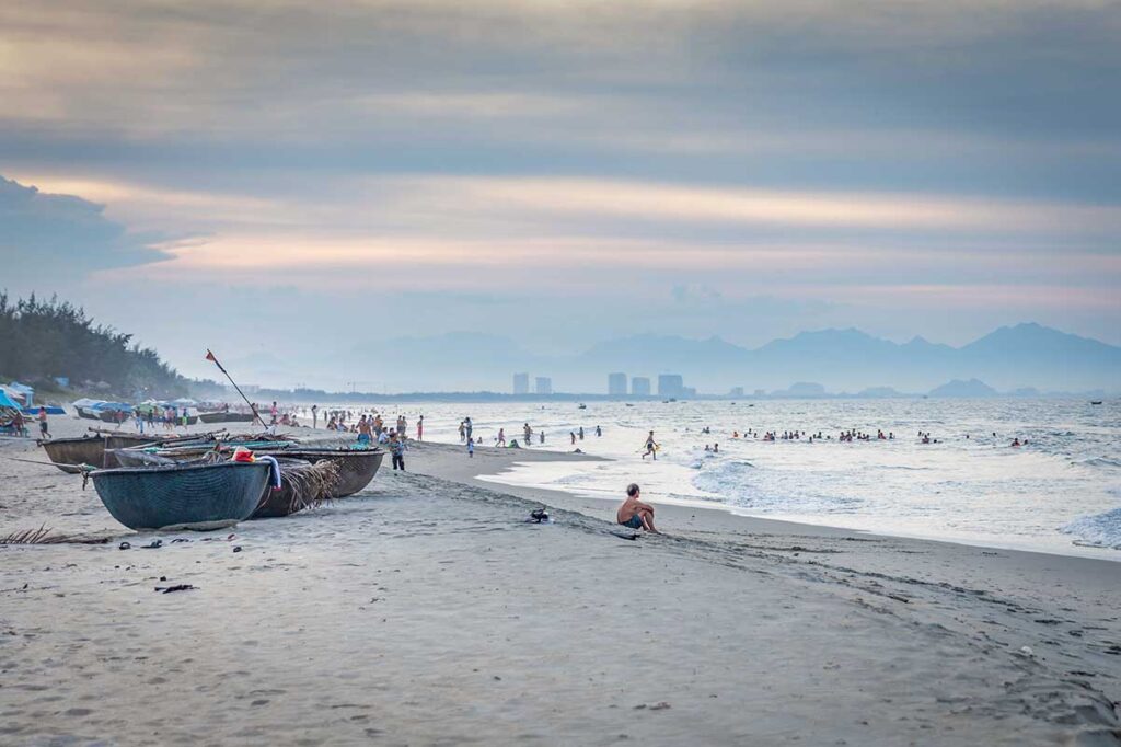 Hoi An Hidden Beach sunset crowd – Local families and travelers enjoy swimming and strolling at Hoi An’s Hidden Beach, with fishing coracle boats on the sand and Da Nang’s skyline faintly visible in the distance.