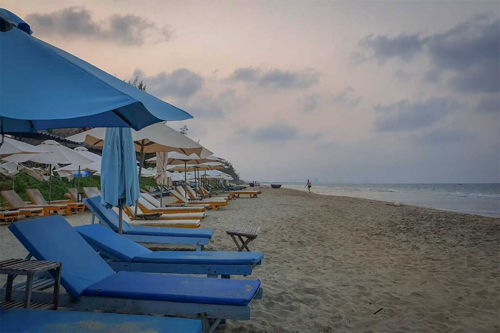 Hoi An Hidden Beach resort sunbeds – Comfortable loungers with white and blue umbrellas lined up along the sandy shoreline of Hidden Beach in Hoi An.