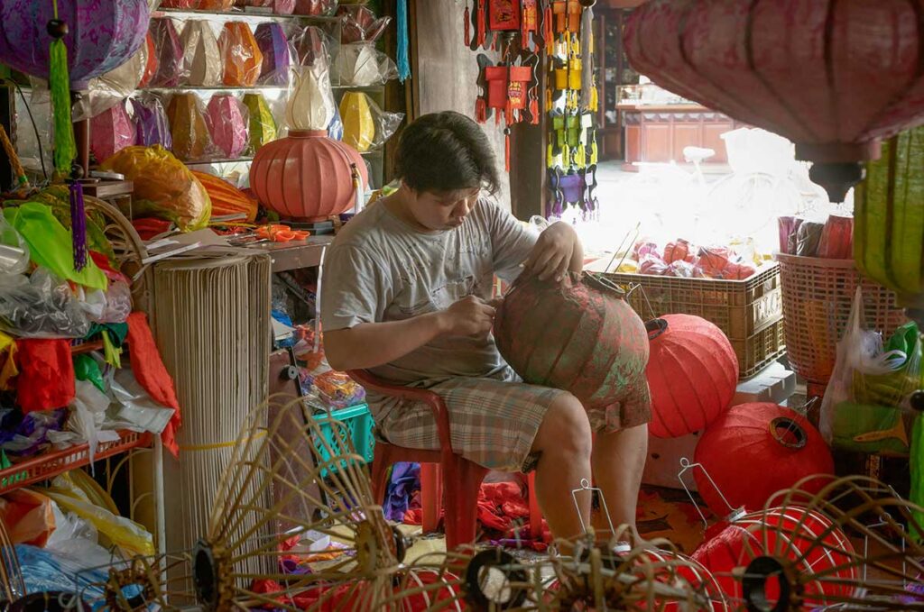 Artisan carefully shaping a silk lantern frame inside a lantern workshop in Hoi An, surrounded by red and colorful handmade lanterns.