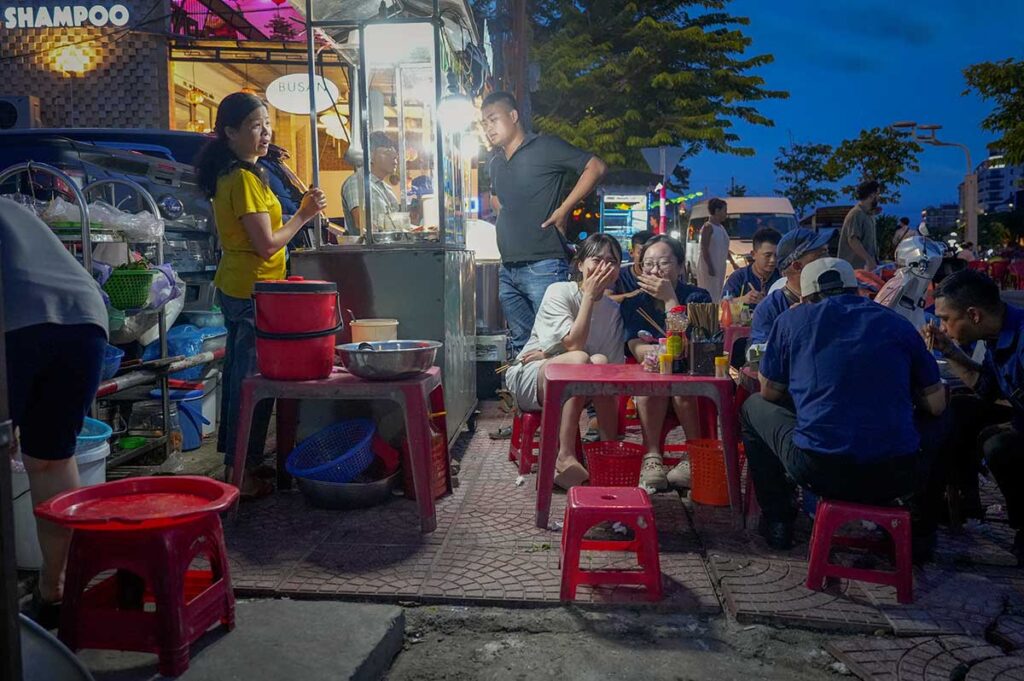 Locals and tourists enjoying street food on small plastic stools at a busy Hoi An night market stall