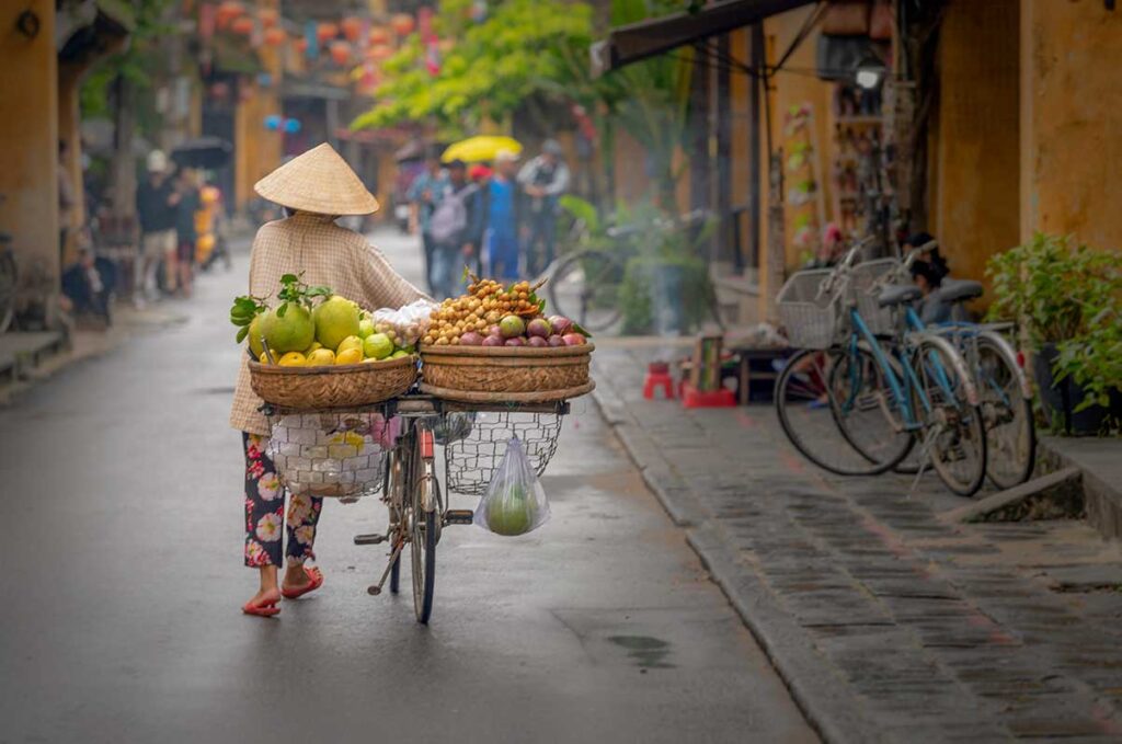 Hoi An street vendor pushing a bicycle with baskets full of tropical fruits like pomelos, rambutans, and passionfruit along the Old Town streets.