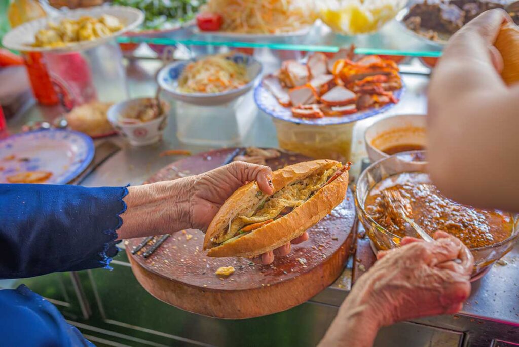 Vendor preparing a famous Hoi An banh mi sandwich with pate, herbs, and fillings at a busy street food stall.