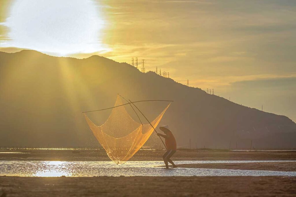 Sunset at Lap An Lagoon were local fishing men is standing in shallow water with a big fishing net