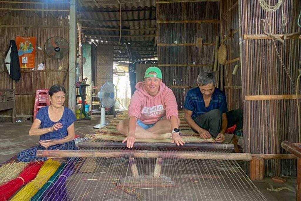 Foreign traveler learning mat weaving beside local artisans in Cam Kim Island’s craft village near Hoi An.