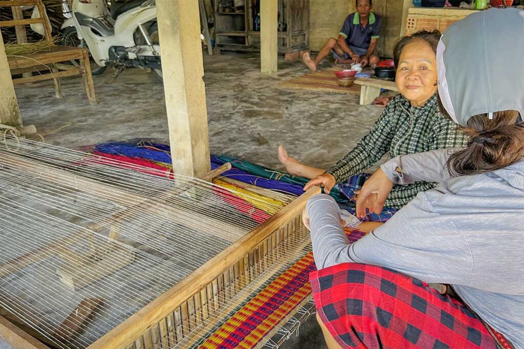 Elderly woman smiling while teaching mat weaving techniques to a visitor inside a traditional workshop on Cam Kim Island.