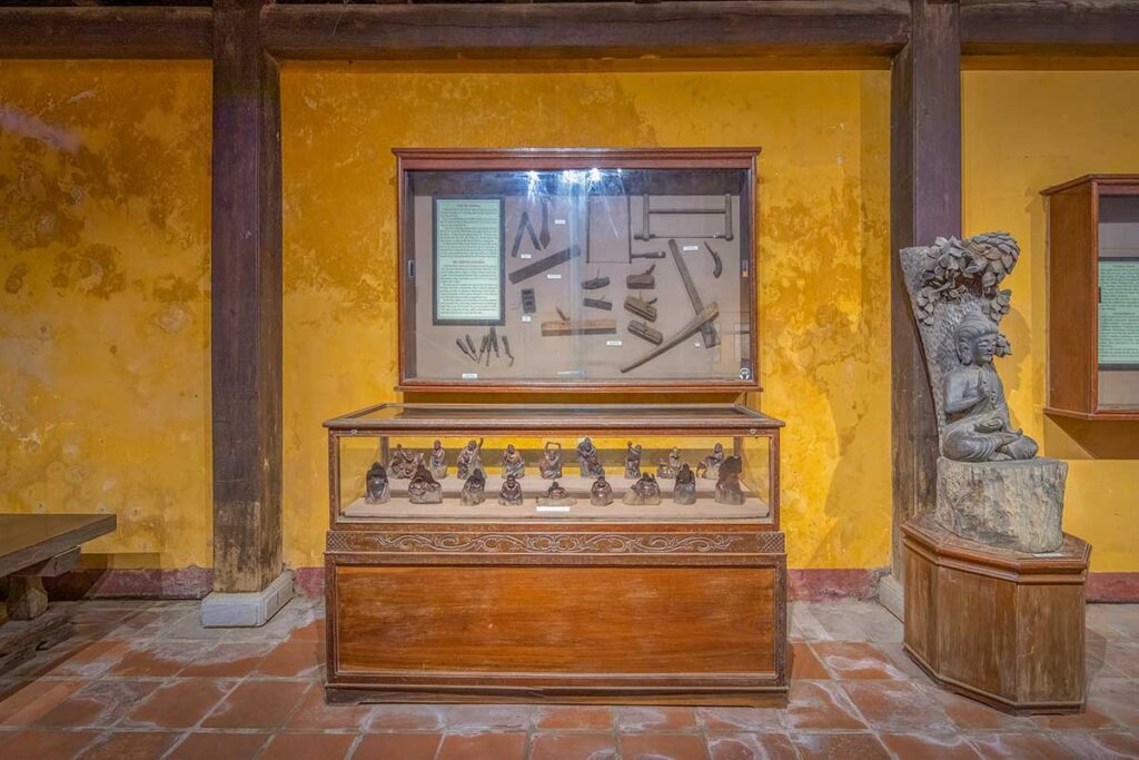 Showcases of carpentry tools and folk statuettes inside the Museum of Folk Culture in Hoi An, next to a carved Buddha sculpture.