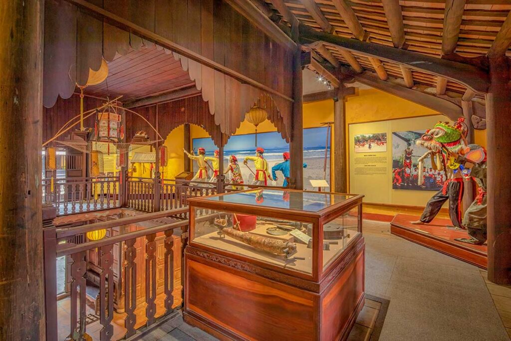 “Performing-arts gallery at the Hoi An Folklore museum featuring festival dioramas, a lion dance figure, and glass cases of traditional instruments.