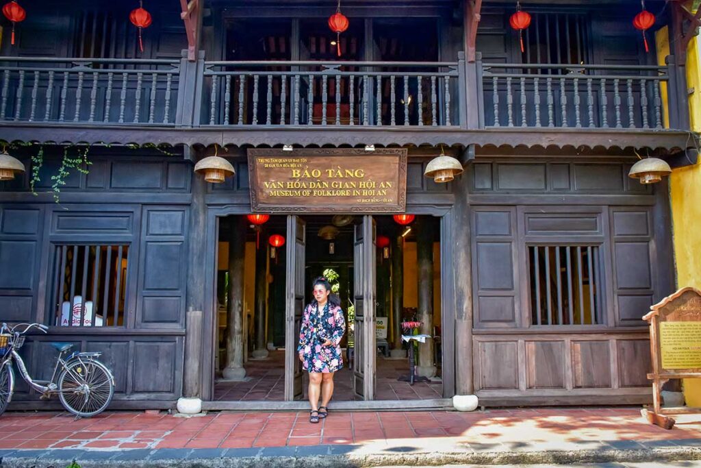 Street-front facade of the Museum of Folk Culture in Hoi An, a heritage timber building on Nguyen Thai Hoc Street with a visitor at the doorway.