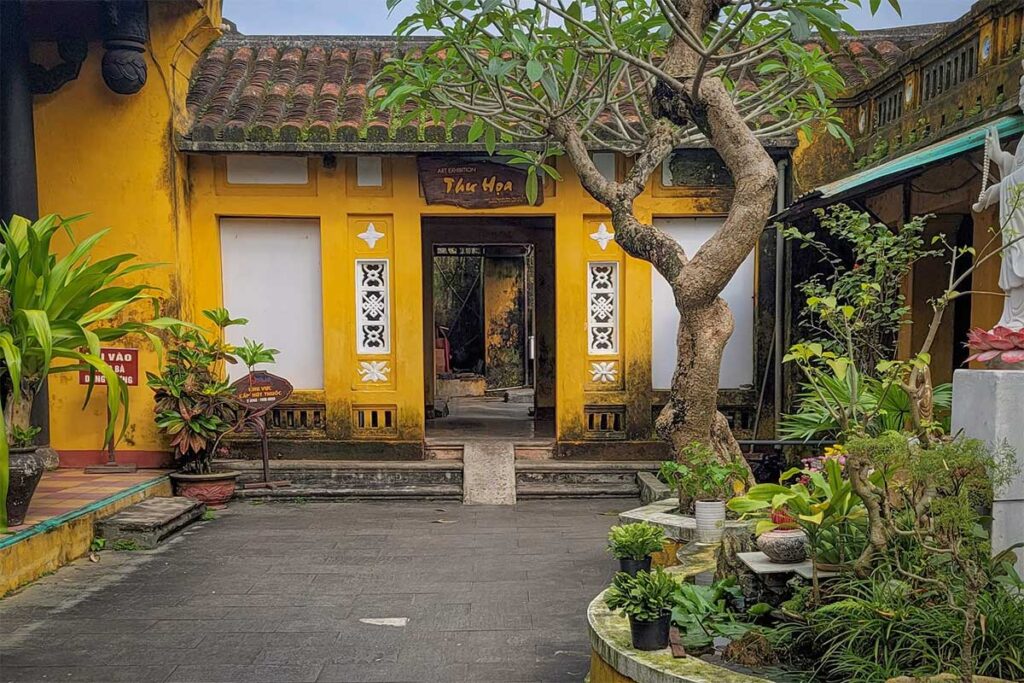 Courtyard view of Quan Am Pagoda Hoi An with bonsai plants, frangipani tree, and historic yellow walls – peaceful atmosphere inside the old temple grounds.