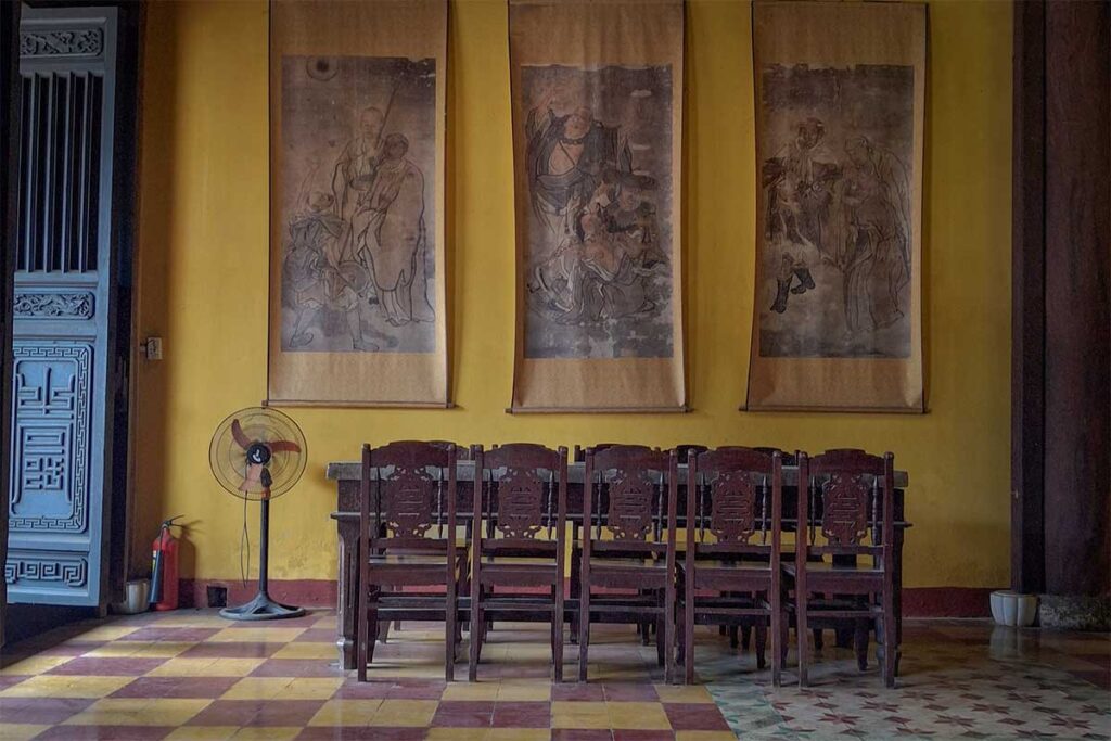 Interior of Quan Am Pagoda in Hoi An with wooden chairs and ancient scroll paintings on yellow walls – highlighting the temple’s historic architecture and cultural artwork.