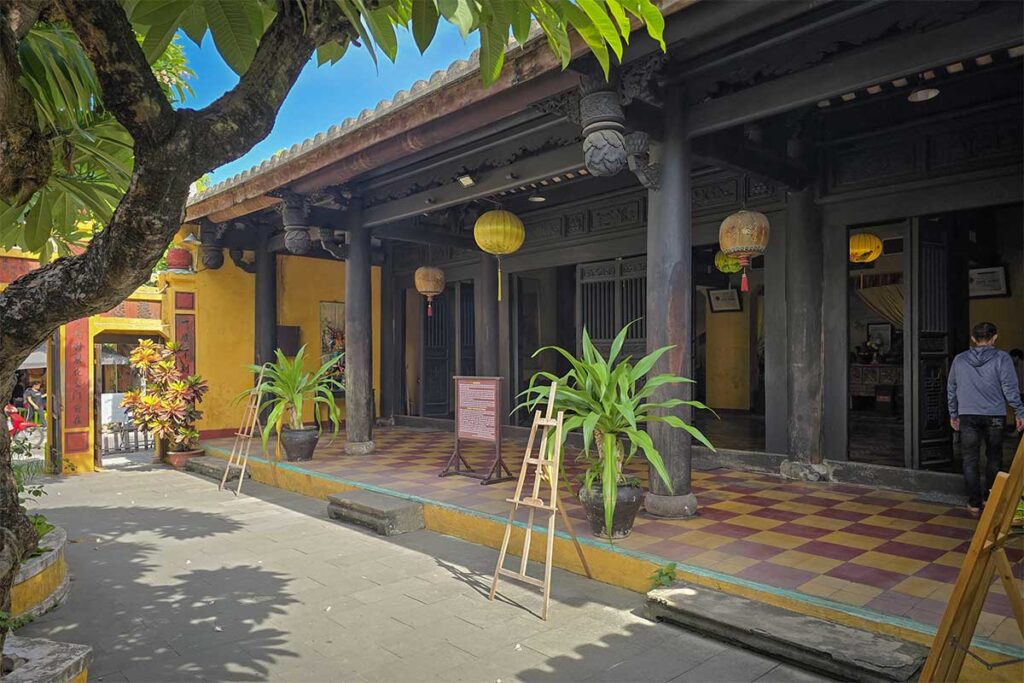 Entrance of Quan Âm Pagoda in Hoi An, with black wooden columns, yellow lanterns, and a shaded walkway under a frangipani tree.